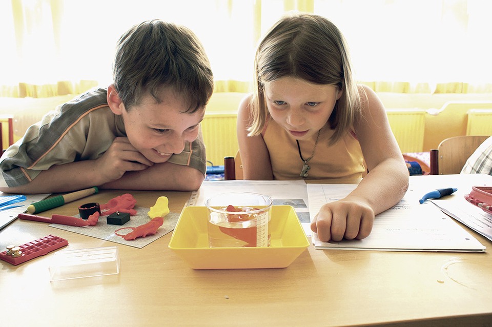 The picture shows two children looking excitedly at an experiment with water in a transparent container. On the table are various craft materials and documents that indicate creative activities.