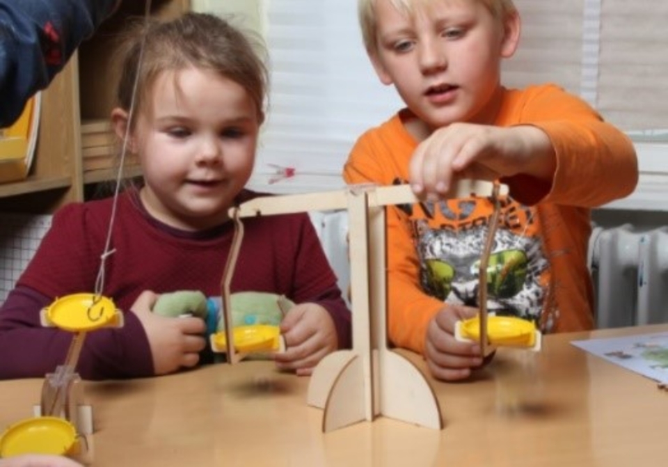 The picture shows two children experimenting on a seesaw to test balance. They each hold a yellow shovel in their hand while they watch with excitement as the seesaw moves.