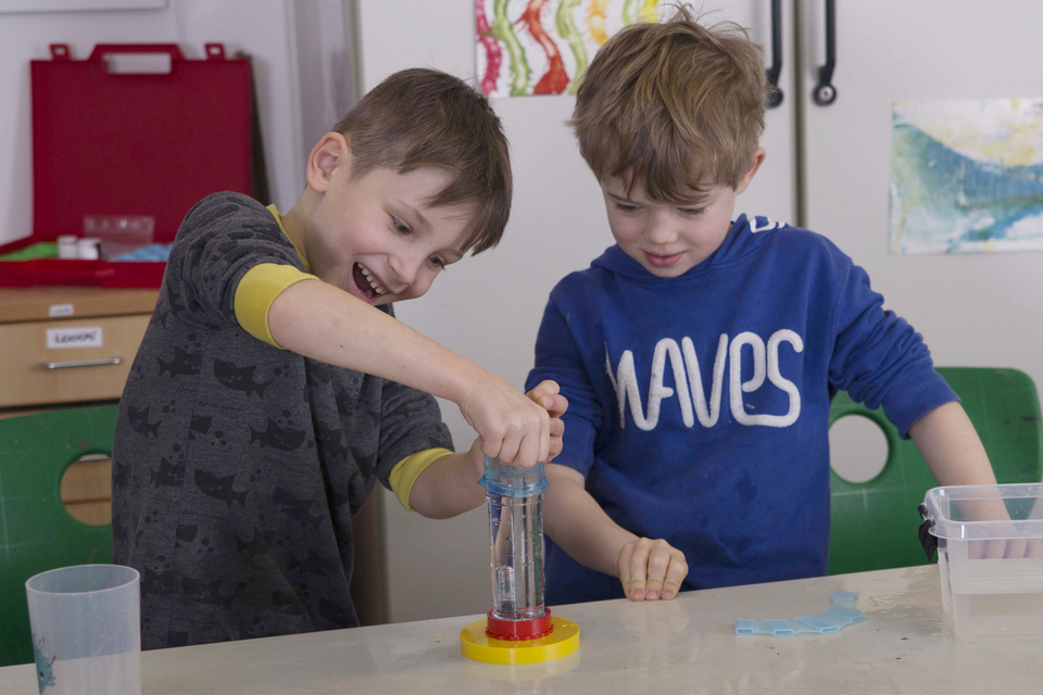 The picture shows two children working together on an experiment. They appear to be manipulating a transparent container filled with water and are clearly excited about the task.