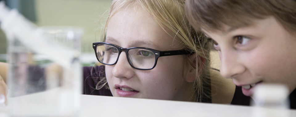 The picture shows two curious children who are staring intently at an experiment in a beaker of water. They appear interested and focused as they follow the experiment&#39;s progress.