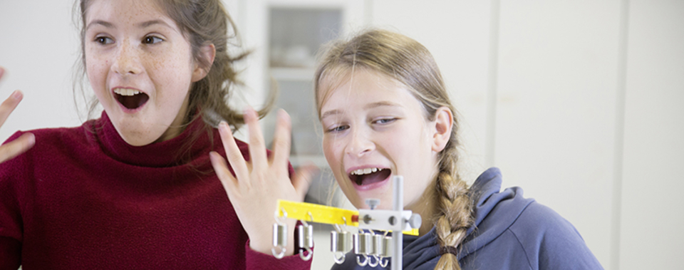 The picture shows two enthusiastically reacting girls standing in front of an experimental device. They show joy and interest in the experiment they are currently observing or conducting.