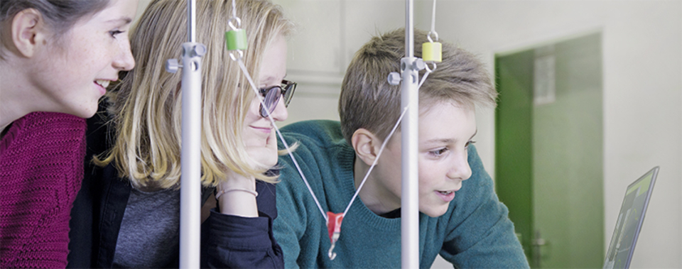 The picture shows three students looking attentively at a laptop and communicating with each other. They seem to be interested in an experiment or scientific task, surrounded by experimental equipment.
