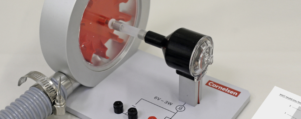 The picture shows an experimental device, a hydroelectric power station for physics lessons. It includes a rotating mechanism and an electrical component, complemented by a circuit that is shown on a labeled worksheet.