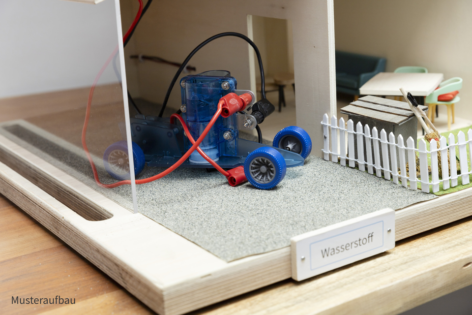 The garage with the fuel cell model car is shown. The car is connected to the roof with a red and black cable.