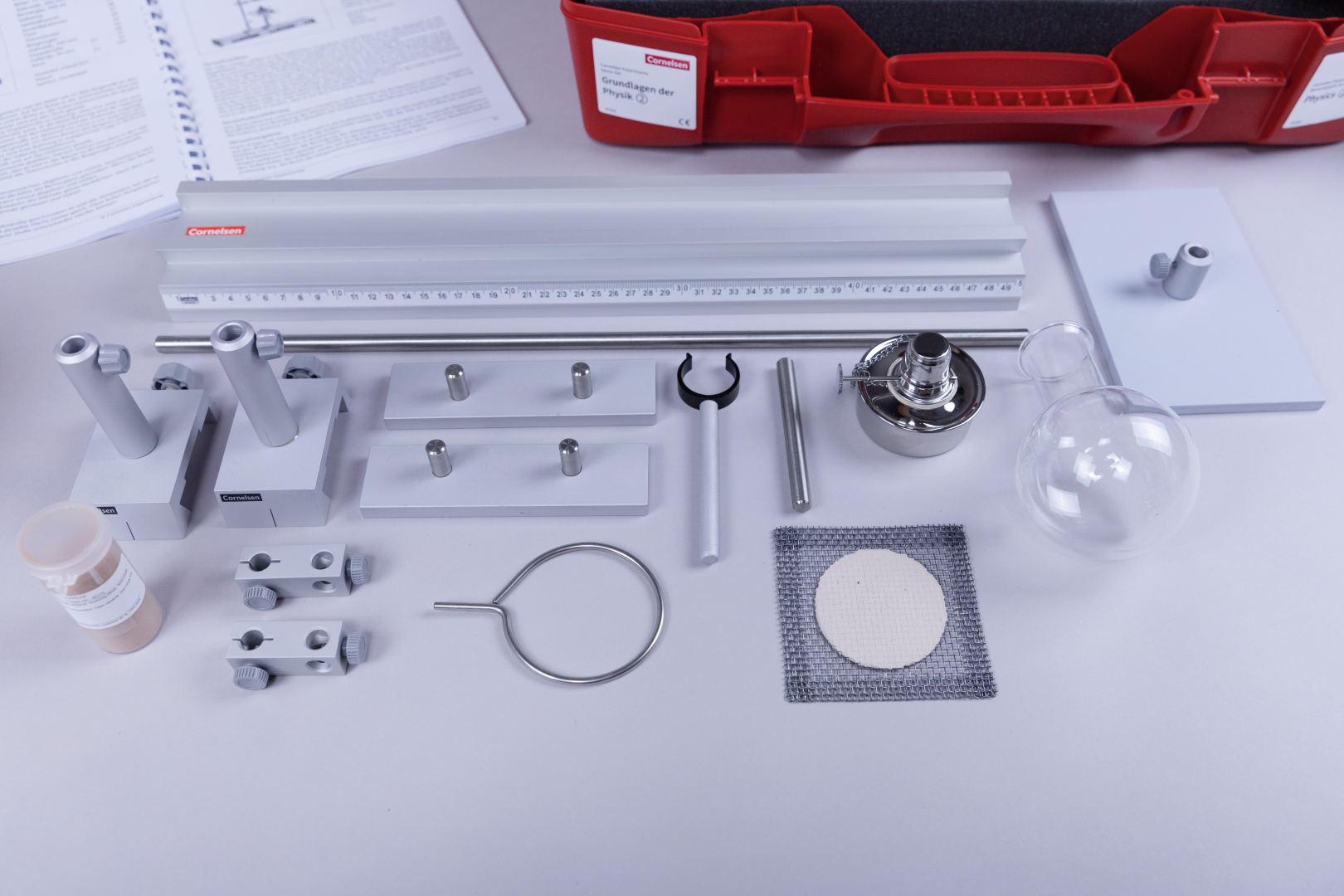 The picture shows a collection of experimental materials, including various holders, a glass bottle, a burner and additional equipment. All these parts are arranged on a table and seem to be intended for experiments in physics classes.