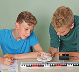 The picture shows two boys participating in an experiment with an optical device. They are working at a table on which an experimental apparatus and worksheets are laid out, while carefully observing and recording the results.