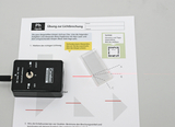 The picture shows an experimental device for light refraction, which consists of a laser module and a glass plate, together with an instruction page. The page contains tasks for investigating the path of light and determining angles of refraction.