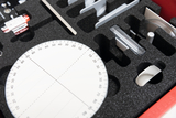The picture shows a set of experimental materials carefully arranged in a foam-padded box. Visible are, among other things, a round measuring circle, various metal parts and a transparent element, which are used for physical experiments.