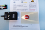 The picture shows an experimental device with laser and LED function, which is positioned on a worksheet for carrying out light experiments. Next to it is a round marker with the logo &quot;Cornelsen Experimenta&quot;, which reflects the light of the laser.