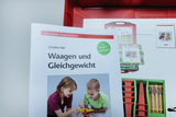 The picture shows a student set entitled &quot;Scales and Balance&quot; by Cornelsen, which contains materials for experiments in education. In the background you can see a red storage box that presents various experimental materials.