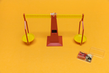 The picture shows a red and yellow seesaw with two yellow bowls that can be used for weight measurements. Next to the seesaw is a transparent container with various small weights and materials for experiments.