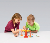 The picture shows two children playing together with an experimental device. They are carefully observing how the weight on a beam balance made of colorful components affects the balance, while various small objects on the table