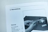 The picture shows instructions for experimenting with heat conduction, in which two glass tubes are held in the hands. In the background, a tea light and a burner frame can be seen on a work surface.