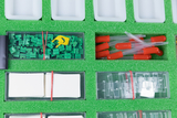 The picture shows a colorful arrangement of experimental materials on green foam. Green components, red pipettes, transparent containers and a small yellow component are visible, all of which can be used for educational purposes.