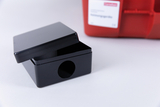 The picture shows a black, square box with a lid and an opening on the side. In the background there is a red container with a label indicating that it is for experimentation.