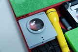 The picture shows an experiment kit with a lens, a yellow flashlight and various other materials suitable for educational and experimental purposes. The kit is placed in a red container with green foam.