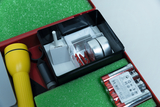 The picture shows an experiment box with various tools and materials, including a flashlight, batteries and other small utensils. The storage unit is designed in a red box with a green foam interior.