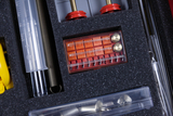 The picture shows a selection of experimental materials in a foam-padded case. These include transparent syringes, metallic components and a special measuring device in a red holder.