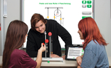 The picture shows three students working on an experiment with pulleys and a pulley. In the background is a digital whiteboard explaining the concepts of fixed and loose pulleys.