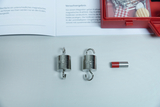 The picture shows magnetic experimental materials, including two weights with hooks and a magnet, as well as a red storage box. In the background, instructions are visible that contain information on experiments with magnetic forces.