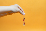 The image shows a hand holding a test tube with two rods, one red and one black. The background is orange, which creates a striking contrast to the transparent glass surface of the test tube.