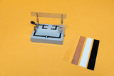 The picture shows an experimental device on an orange surface that is used to carry out vibration experiments. Next to the device are various materials in the form of strips that can be used for the experiments.