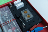 The picture shows a set of experimental materials that includes various components such as clamps, cables and transparent containers with gold connectors. It is organized in a red box with a textured foam pad to support the te