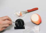 The picture shows a hand working with tweezers on a special cutting device, while a halved apple and a knife are lying next to it. In addition, cover glasses and a bowl with apple pieces are arranged on a table surface, suggesting a