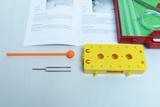 The picture shows an experiment kit with a yellow circuit block, a tuning fork, a red rod and an orange ball. Next to it is an instruction manual that contains the materials and procedures for various experiments in the educational field.