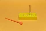 The picture shows a yellow experimental device with several holes in which a tuning fork is inserted. Next to the device is an orange plastic rod, probably for conducting experiments in the educational field.