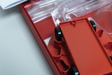 The picture shows a red experiment box containing a vehicle kit. A red vehicle part with black wheels is visible, while transparent plastic parts and instructions are in the background.