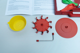 The image shows various components of an experiment kit, including a yellow container, a red rotating part and a metal tube. In the background are instructions for conducting experiments dealing with mecha