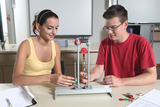 The picture shows two students working on an experimental device used for physics or technology lessons in education. The boy and the girl interact with various weights and a suspension system while paying attention