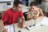 The image shows two students working on an experimental device to explore physics concepts. The focus is on an apparatus with weights and a measuring cylinder, while both carefully take notes and discuss.