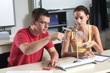 The picture shows two students experimenting with a scale at a table. The boy is lifting a measuring block while the girl is holding a small plate on the other side of the scale to compare the weight.