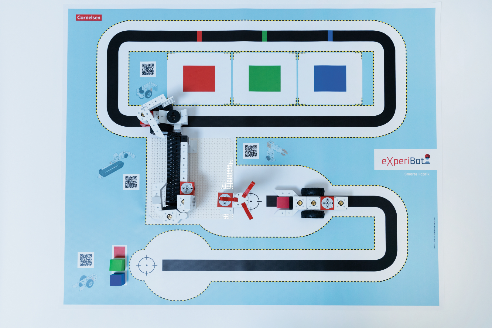 The picture shows an experimental platform with a rotating robot and a colored track for programming and testing. Next to the track there are various QR codes and colored elements that can be used for interactive learning activities.