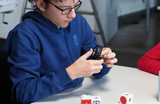 A boy wearing glasses examines a small rubber wheel while sitting at a table. In front of him are various building blocks in white and red, possibly intended for an experiment or an educational project.