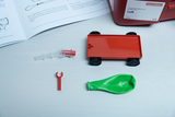 The picture shows various experimental materials, including a red vehicle with wheels, a green balloon and other components that can be used for experimental purposes. In the background there is an instruction manual that explains the use of these parts.