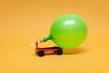 The picture shows a small vehicle with a green balloon mounted on it. The vehicle is standing on an orange surface and is probably demonstrating a principle of air pressure or kinetics.