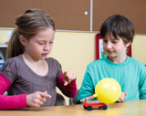 The picture shows two children working on an experiment with a small, red-painted vehicle equipped with a yellow balloon. The children seem interested and engaged in the experiment while they are using the mechanics