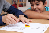 The picture shows two children actively working on an experimental project. One boy is lifting a magnet above a piece of paper with a road drawn on it, while the other watches intently, with a small yellow vehicle placed in the center.