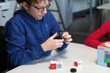 The picture shows a student holding a black element in his hand and combining it with a construction toy on the table. Several colorful modules lie on the table while another student in a red shirt observes.