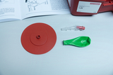 The picture shows an experiment kit for educational institutions, consisting of a red base plate, a green balloon and a connecting part. Next to the materials is a manual containing instructions on how to carry out an air experiment.