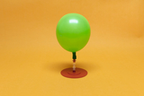 The image shows a green balloon decoration mounted on a sturdy, round red base. The background is a soft yellow, which highlights the vibrant color of the balloon and creates a friendly atmosphere.