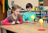 The picture shows two children looking intently at two yellow balloons that are on a table. The balloons are connected by transparent tubes and the children appear to be taking part in an experiment that teaches scientific concepts.