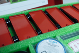 The picture shows a set of red plastic parts with wheels arranged in a green foam insert. Next to these components, various experimental materials such as Vaseline and measuring instruments can be seen, which are intended for educational purposes.