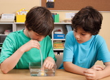 The picture shows two boys working in an experiment kit. One of the boys is using a tube to move a small yellow ball in a container of water while the other boy watches attentively.