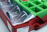 The picture shows a red box containing various laboratory equipment, including several clear funnels and beakers, safely padded in green foam. This equipment is specifically designed for experiments in German educational