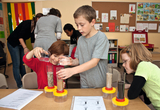 In the picture, children in a classroom are experimenting with different materials in clear containers. They add water to the containers and observe the results, with their curiosity and cooperation clearly visible.