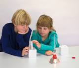 The picture shows two children sitting at an experiment station and working concentratedly with various laboratory equipment. A red-coloured liquid in an Erlenmeyer flask is rising in the foreground, while the children are holding a pipette and a measuring cylinder 